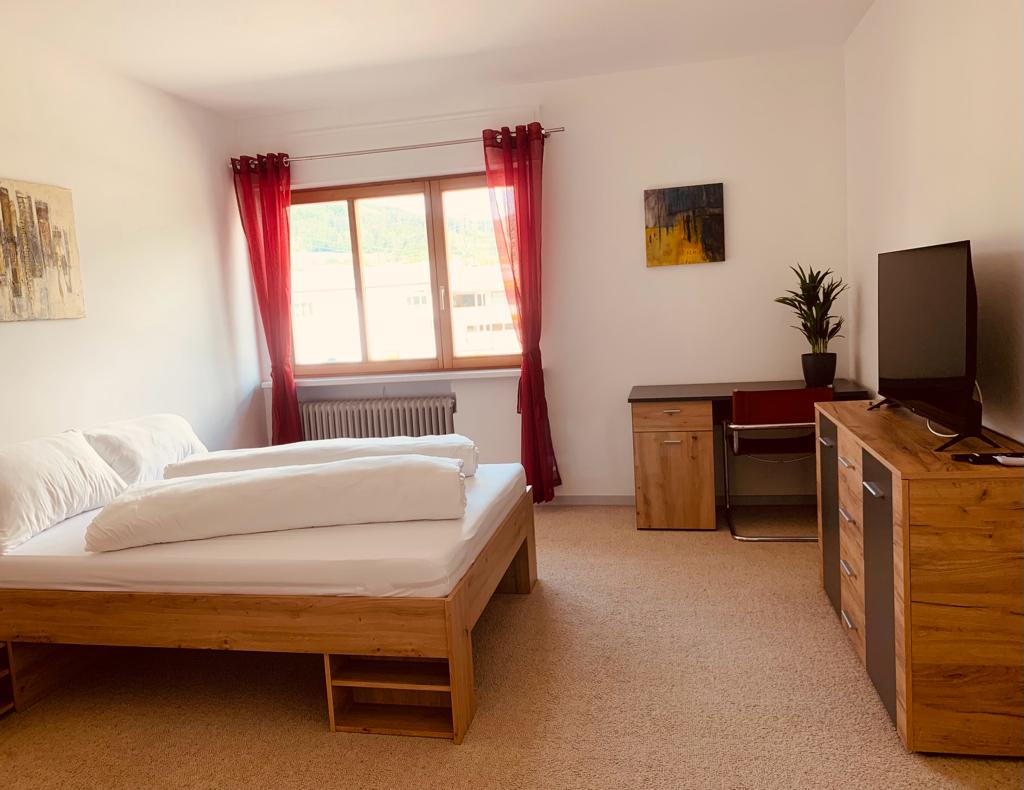 A well-lit bedroom features a spacious bed with a white comforter and decorative pillows. A wooden desk and chair are positioned next to a TV stand, which holds a modern television. Natural light enters through a large window adorned with red curtains.