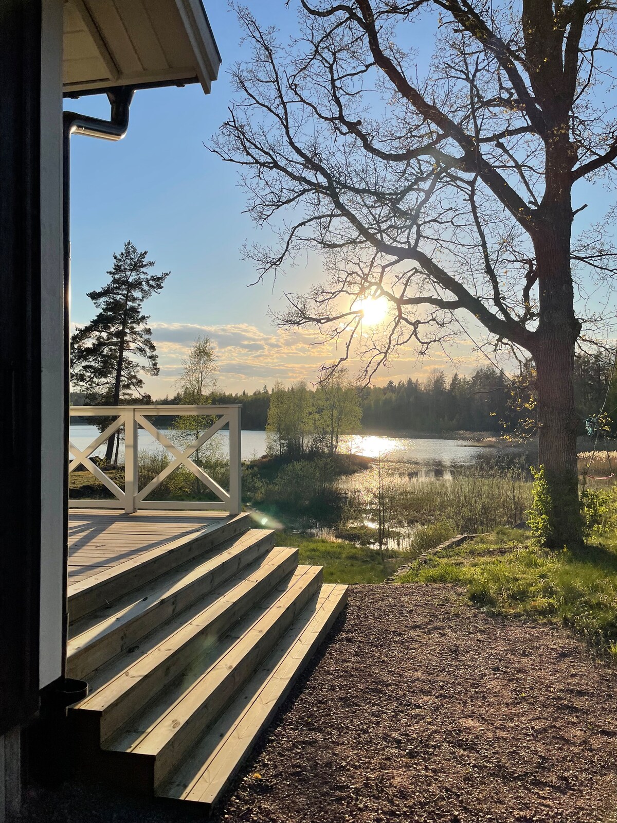Steps lead down from a wooden deck, framed by a peaceful lakeside scene. Sunlight glimmers on the water's surface, while trees surround the area, providing a natural setting that invites relaxation.