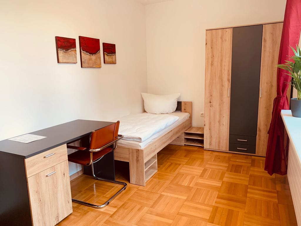 A well-furnished single bedroom features a spacious bed with a white comforter. A desk with a chair is positioned next to the bed, and a wardrobe is visible against the wall. Three abstract paintings adorn the wall, adding a touch of color to the room.