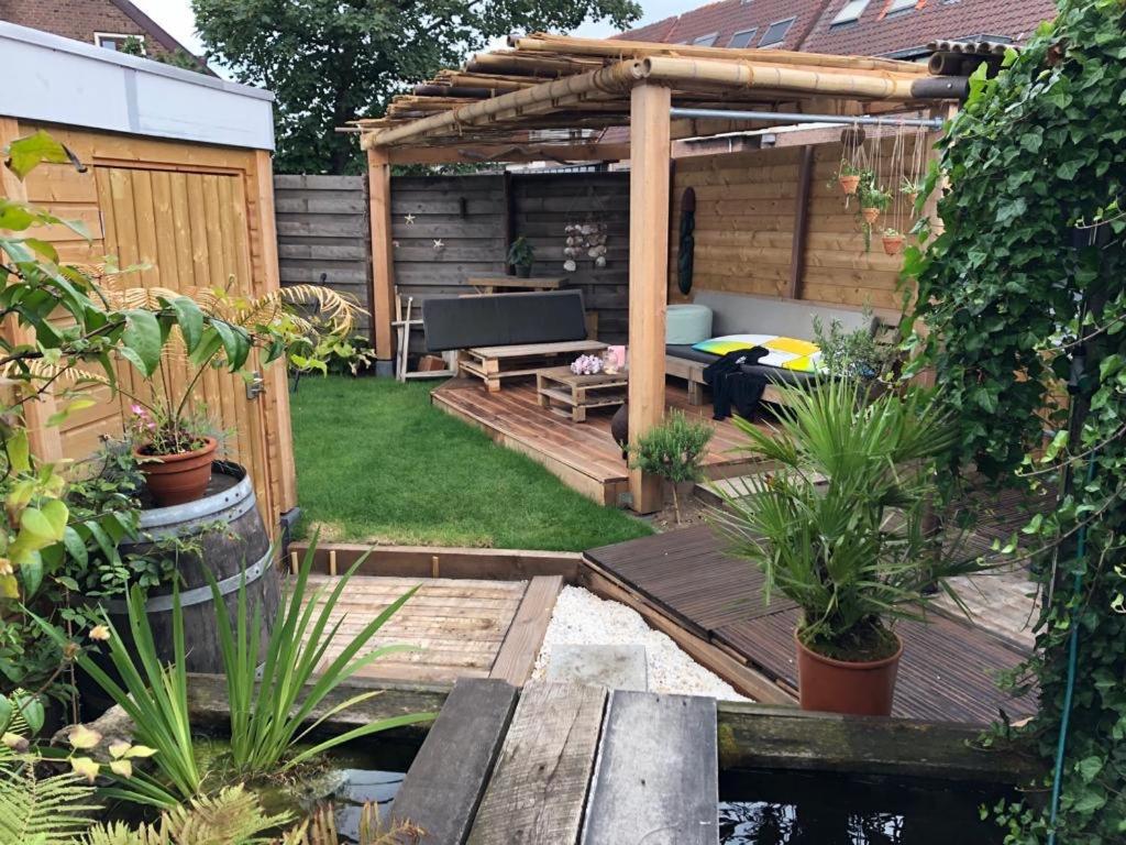 A tranquil tropical garden is showcased, featuring a wooden deck and lounging area surrounded by greenery. A small pond and barrel planter add to the serene atmosphere. Comfortable seating is available, with an inviting color palette enhancing the space.