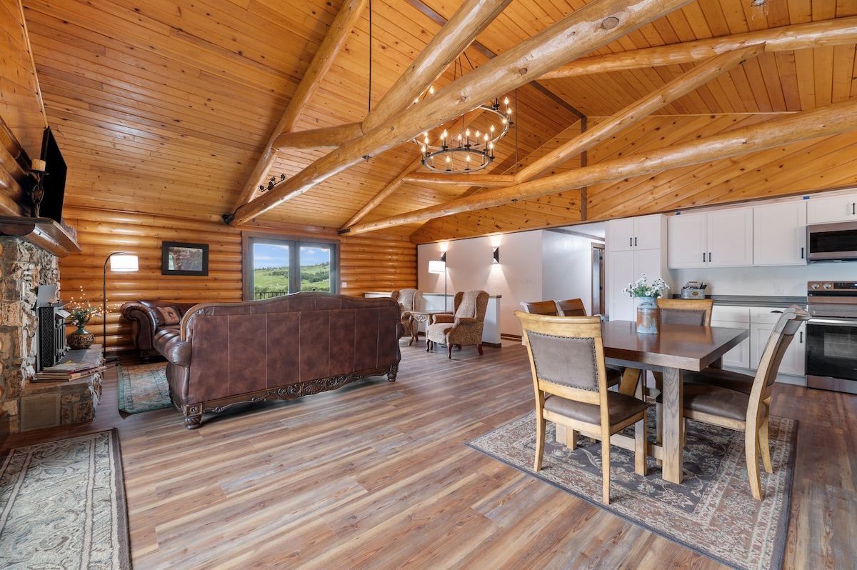 A spacious living area features high wooden ceilings with exposed beams. Comfortable rustic seating and dining arrangements are visible, complemented by a stone fireplace. Large windows provide views of the surrounding landscape, enhancing the open floor concept that encourages connection among guests.