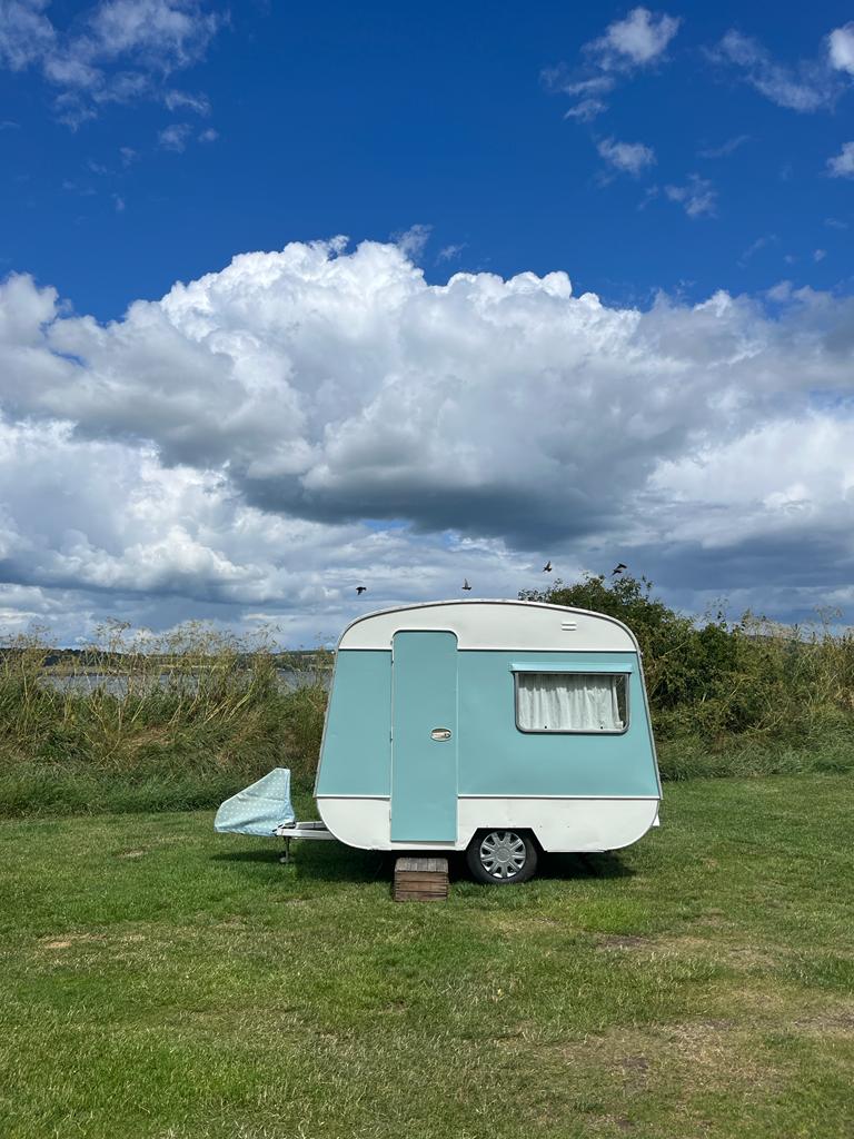 A vintage caravan with a soft blue exterior is parked on lush green grass beside a calm waterway. A small beach shelter is positioned nearby, with fluffy white clouds scattered across a bright blue sky.