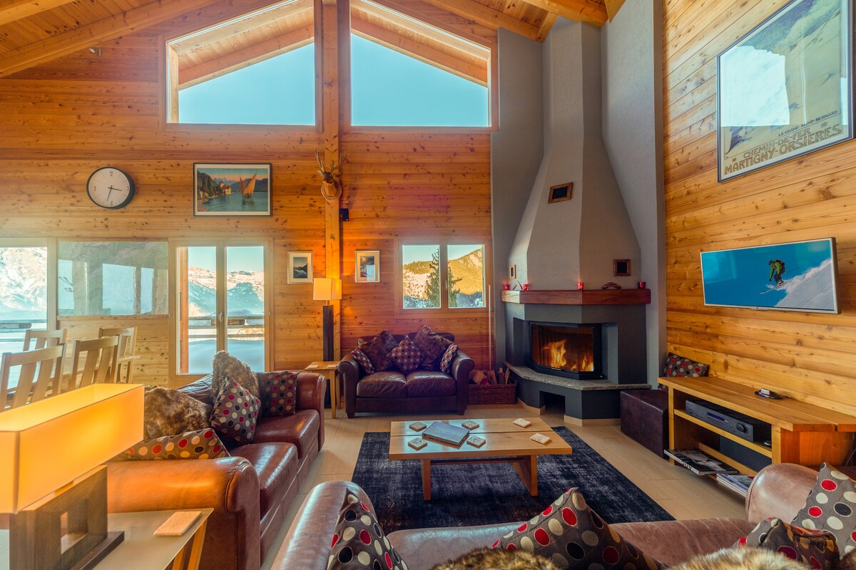 A welcoming living area features wooden walls and large windows that provide natural light and mountain views. Leather sofas are arranged around a central coffee table, with a fireplace set against a backdrop of warm wood. Artwork is displayed on the walls, adding visual interest.