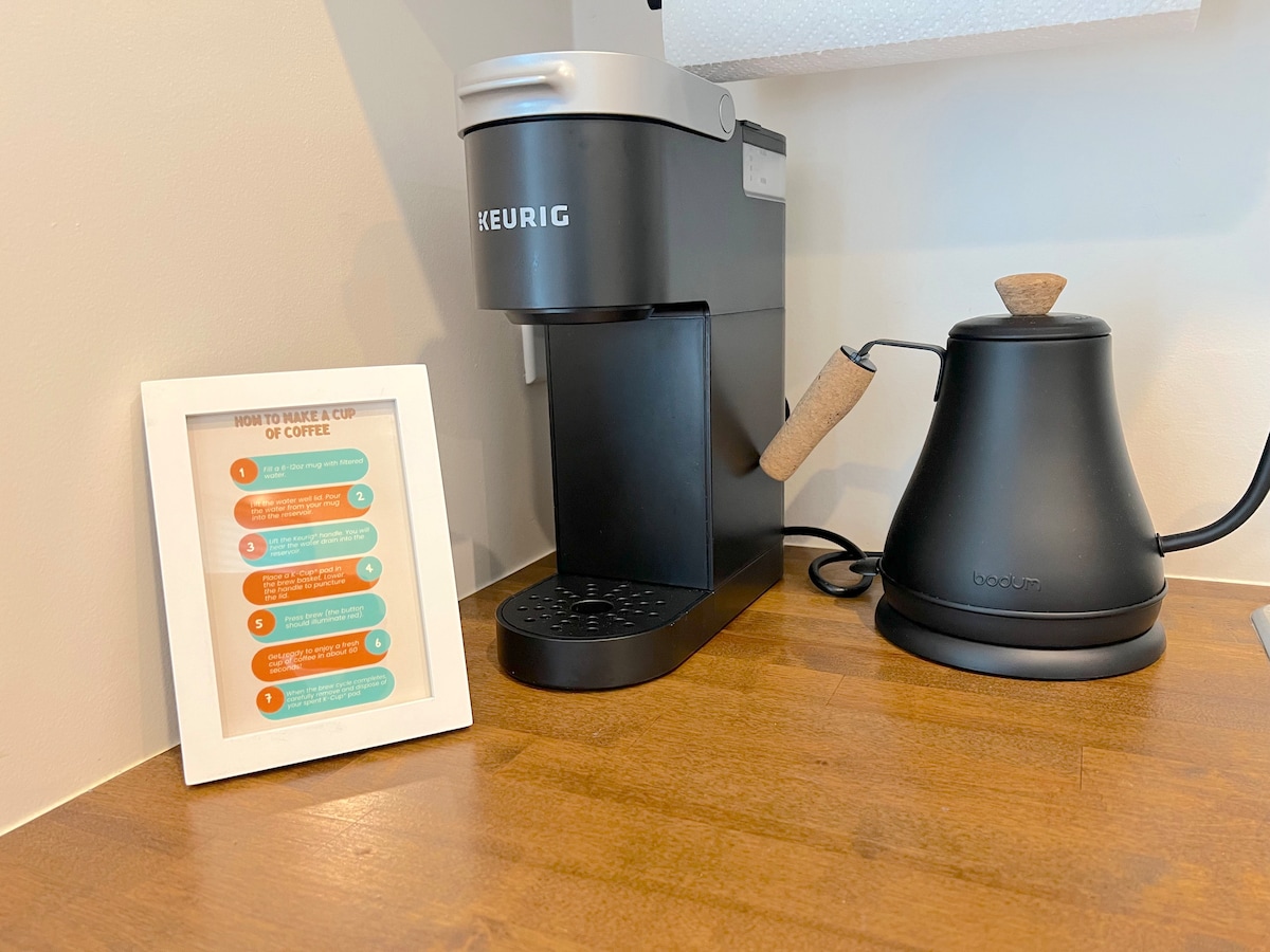 A coffee station features a Keurig machine alongside a sleek black kettle. A framed instruction guide for brewing coffee is placed nearby, providing clear steps. The warm wooden surface complements the modern design, creating a functional setup for guests.
