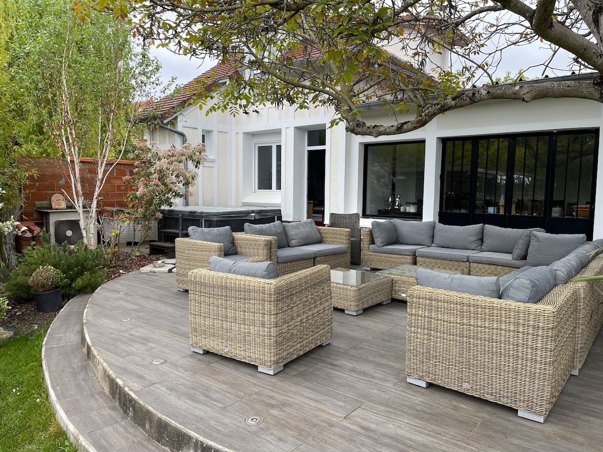 An outdoor seating area is presented, featuring a circular arrangement of wicker furniture with plush gray cushions. A large wooden deck supports the seating, while greenery and trees provide a natural backdrop, enhancing the inviting outdoor atmosphere.