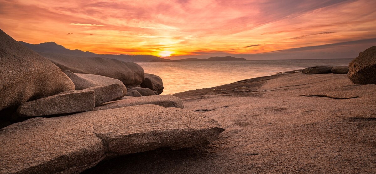 A coastal scene showcases smooth, large rocks leading to the water's edge, with the sun setting on the horizon. The sky is painted in soft hues of orange and pink, reflecting on the calm surface of the sea in the distance.