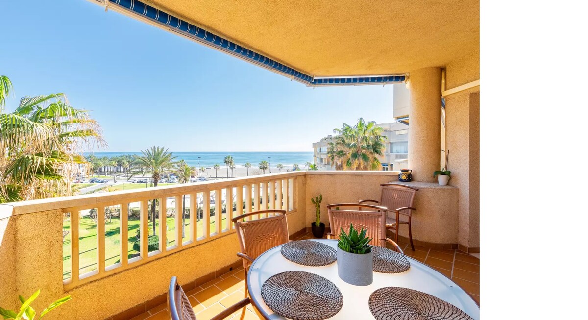 A spacious terrace features a dining table surrounded by four chairs, offering views of the sea and gardens below. Potted plants add a touch of greenery, while the clear blue sky enhances the bright and inviting atmosphere.