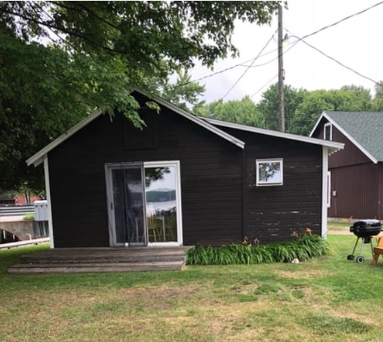 The exterior of the cabin features dark wooden siding, with a large sliding glass door and a small window providing a view of the surrounding area. A charcoal grill is located on a grassy patch in front, with colorful flowers along the base.