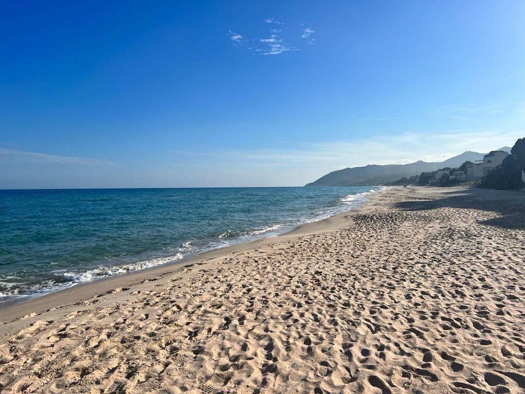 A wide expanse of sandy beach stretches along the shoreline, meeting gently lapping waves of a calm sea. The coastline is bordered by distant hills, with a clear blue sky overhead, enhancing the tranquil setting.
