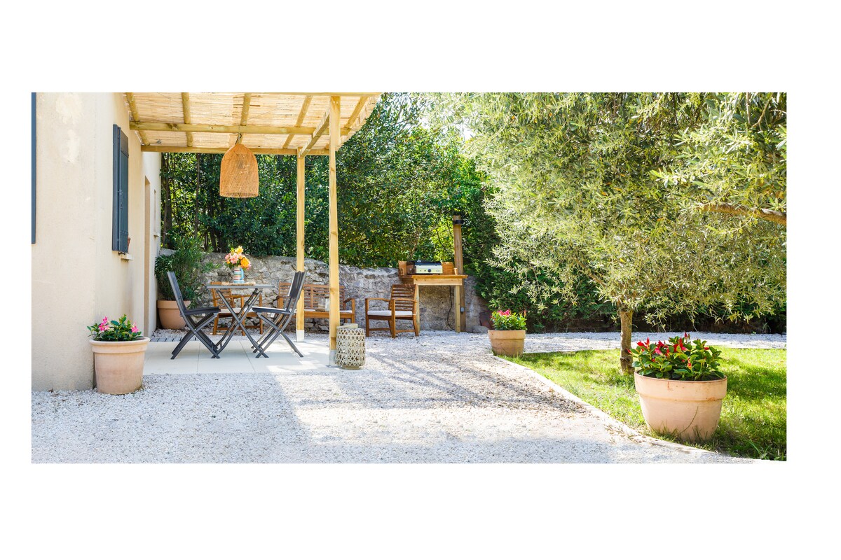 An outdoor terrace is set with a wooden table and four chairs, surrounded by greenery. Potted plants add color to the landscape, while a barbecue area is visible in the background. Natural light highlights the inviting space under a shaded pergola.