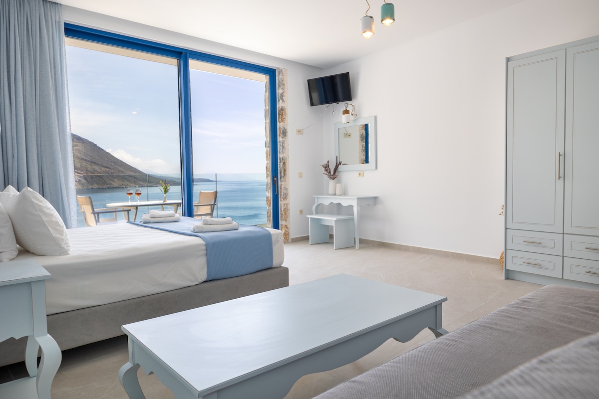 A bright room showcases large windows with stunning sea views, allowing natural light to fill the space. A spacious bed is neatly made with light linens. A small table and chairs are positioned by the window, providing a spot for relaxation. A flat-screen television is mounted on the wall.