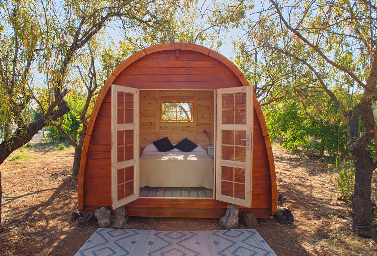 A wooden cabin is seen nestled among trees, featuring wide, welcoming doors that open to a cozy interior. A spacious bed is visible, complemented by natural light streaming through a small window. A patterned rug is situated outside the entrance, enhancing the serene outdoor setting.