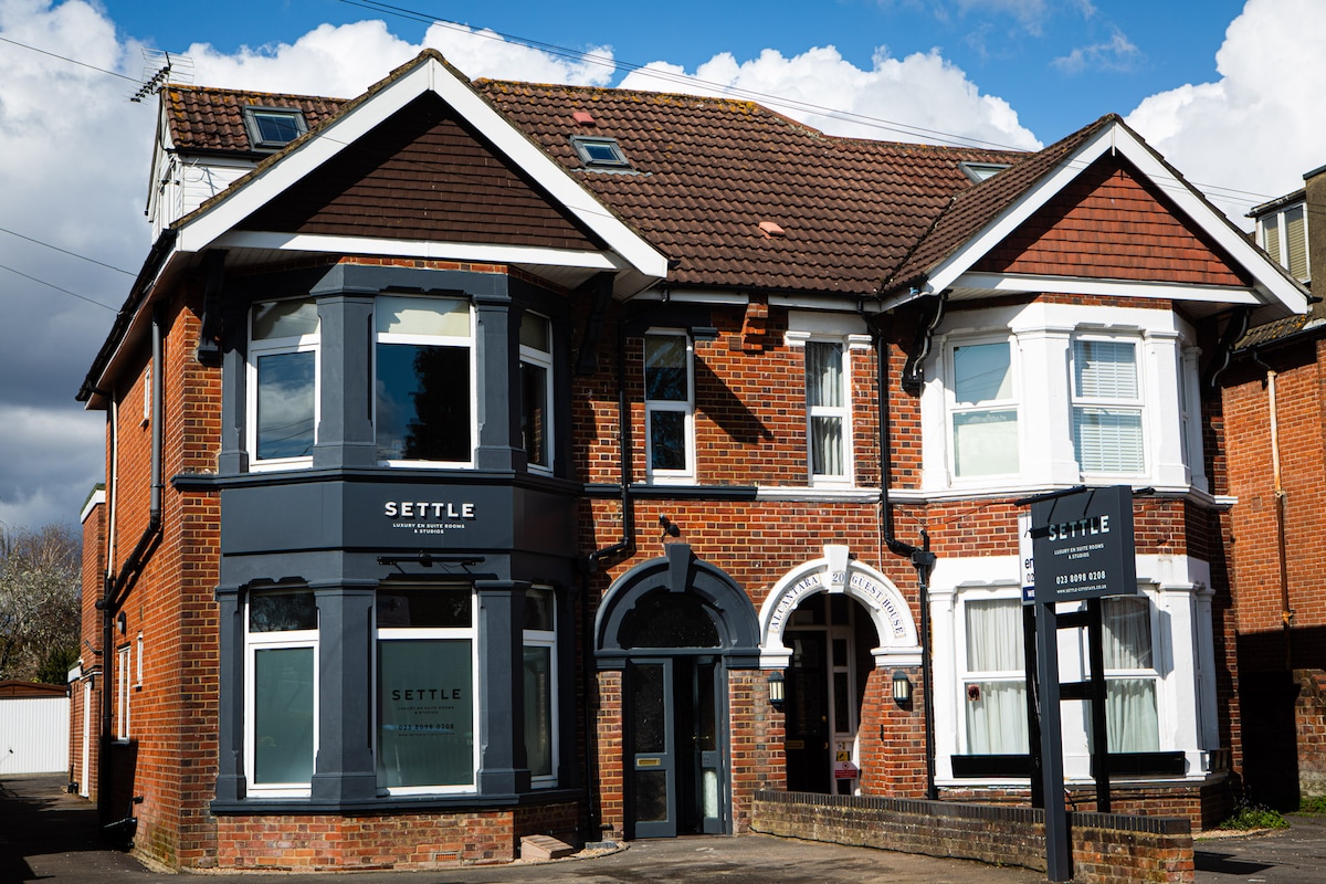 The exterior of a boutique hotel features a classic brick facade, displaying large windows and a welcoming entrance. A sign reading 'SETTLE' is visible, while modern architectural elements complement the traditional design. The surrounding area is landscaped, contributing to the inviting appearance.