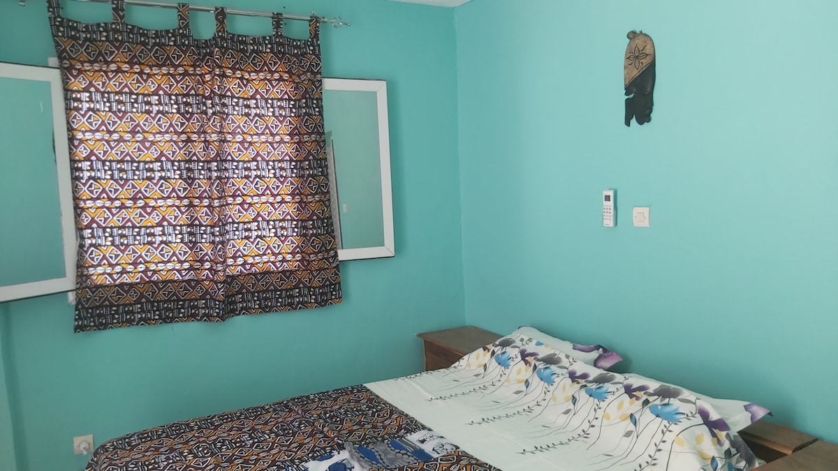 A sleeping area features a spacious bed adorned with patterned bedding. Bright turquoise walls complement the intricate window curtains. A wooden bedside table is positioned next to the bed, with a decorative wall hanging above, contributing to the inviting environment.