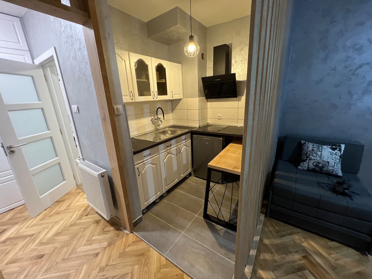 A compact kitchen area is presented, featuring white cabinetry and a contrasting wooden countertop. A modern range hood is mounted above the stove, while a cozy seating area is visible to the right, complemented by a soft sofa and decorative pillows.