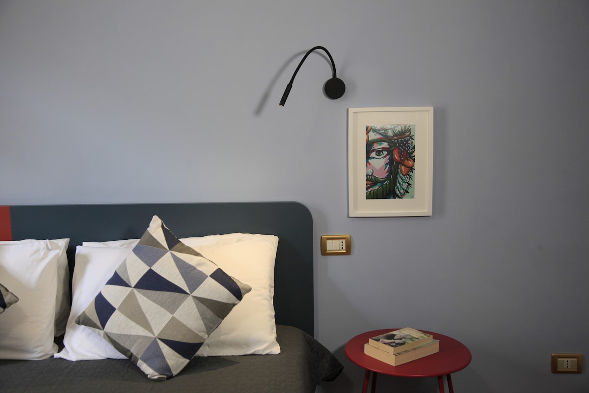 A cozy sleeping area is highlighted by a bed with patterned pillows. A small red side table is positioned beside the bed, adorned with books. A colorful piece of wall art is displayed above the bed, complemented by a modern black wall sconce.