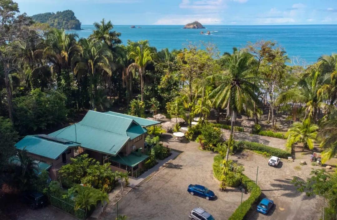 An aerial view captures the property surrounded by lush tropical vegetation and a sandy beach in the background. The spacious parking area is visible, along with an inviting green-roofed building nestled among the trees, providing a serene coastal setting.