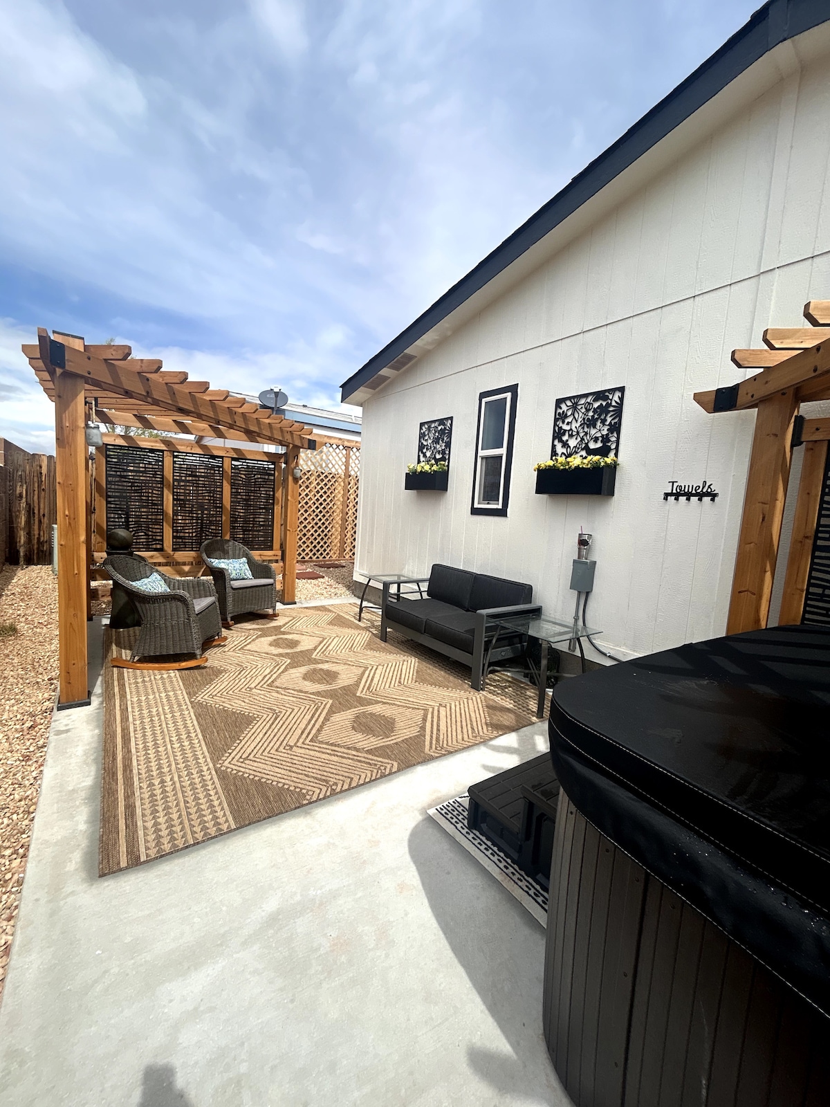An outdoor patio area features comfortable seating under a wooden pergola, with two accent chairs and a loveseat arranged over a patterned rug. Potted plants adorn the wall above, and a hot tub is positioned nearby, contributing to a relaxing outdoor environment.