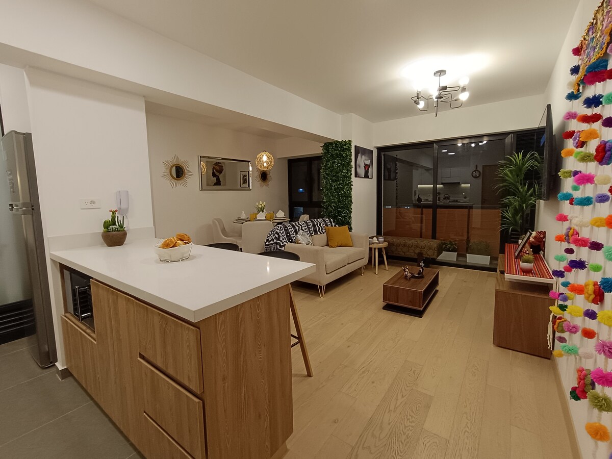 A modern living space showcases a spacious layout. An open kitchen flows into the living area, which features a cozy seating arrangement with a small coffee table. Bright natural light filters through large windows, while a colorful decorative wall adds a playful touch.