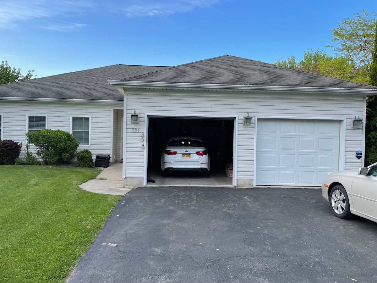 A single-story home is shown with a neatly manicured lawn and a two-car garage. One vehicle is parked inside the garage, while another is positioned in front of it. The exterior features light-colored siding and a simple roofline under a clear blue sky.