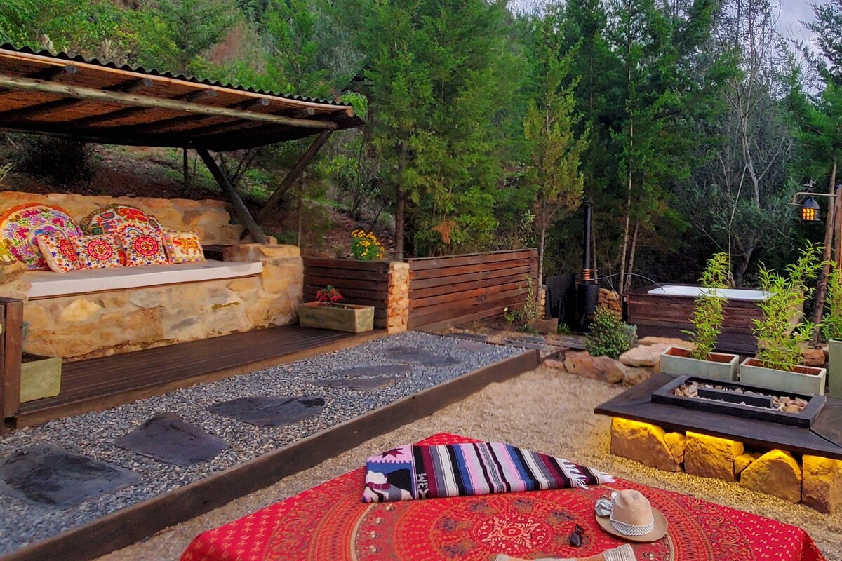 An outdoor gathering space features a wooden structure with a comfortable seating area adorned with colorful cushions. Nearby, a fire pit is surrounded by smooth stones, with lush greenery providing a natural backdrop. A decorative tablecloth is placed on a low table, enhancing the tranquil setting.