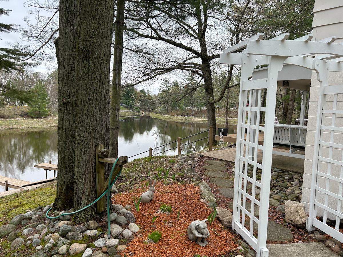 A serene view of a pond is framed by trees and a white lattice structure. Decorative stones and natural landscaping enhance the area, with small garden statues positioned near the water's edge. The setting offers a peaceful retreat surrounded by nature.