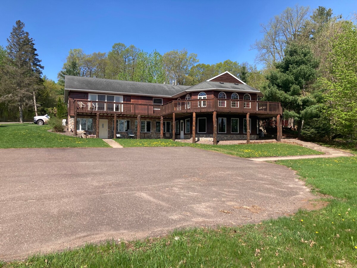 A spacious two-story exterior features a mix of wood and stone detailing. Large windows allow natural light to fill the building. A broad deck with railings extends along the front, surrounded by trees and green grass, leading to a paved parking area.