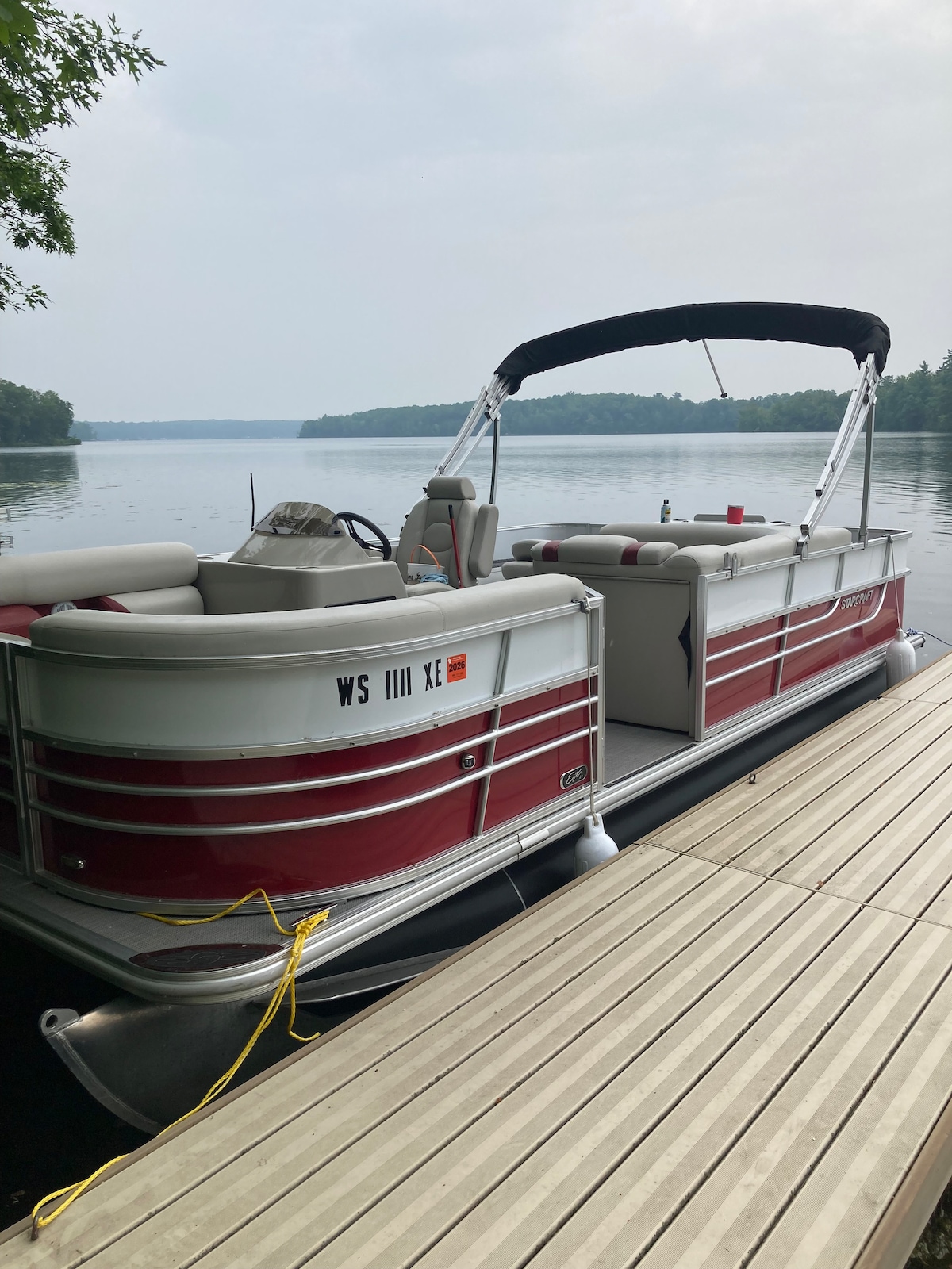 A pontoon boat is docked along a wooden pier, facing calm waters. The boat features cushioned seating in light grey and red tones, with a canopy offering shade. Lush greenery surrounds the serene lake, contributing to a tranquil environment.