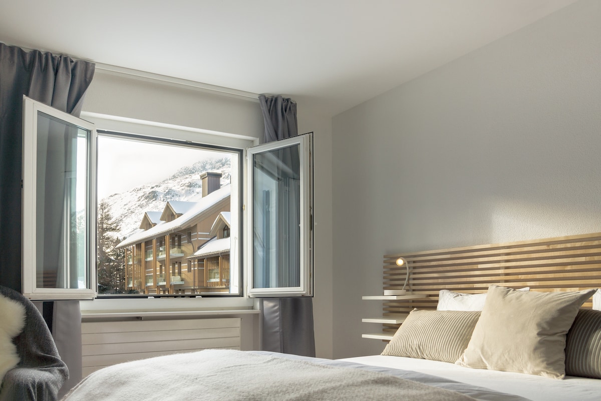 A spacious bedroom features large windows that open to reveal a snowy mountain landscape outside. The bed is adorned with neutral-toned pillows and a cozy blanket. Soft curtains frame the windows, allowing natural light to brighten the room.