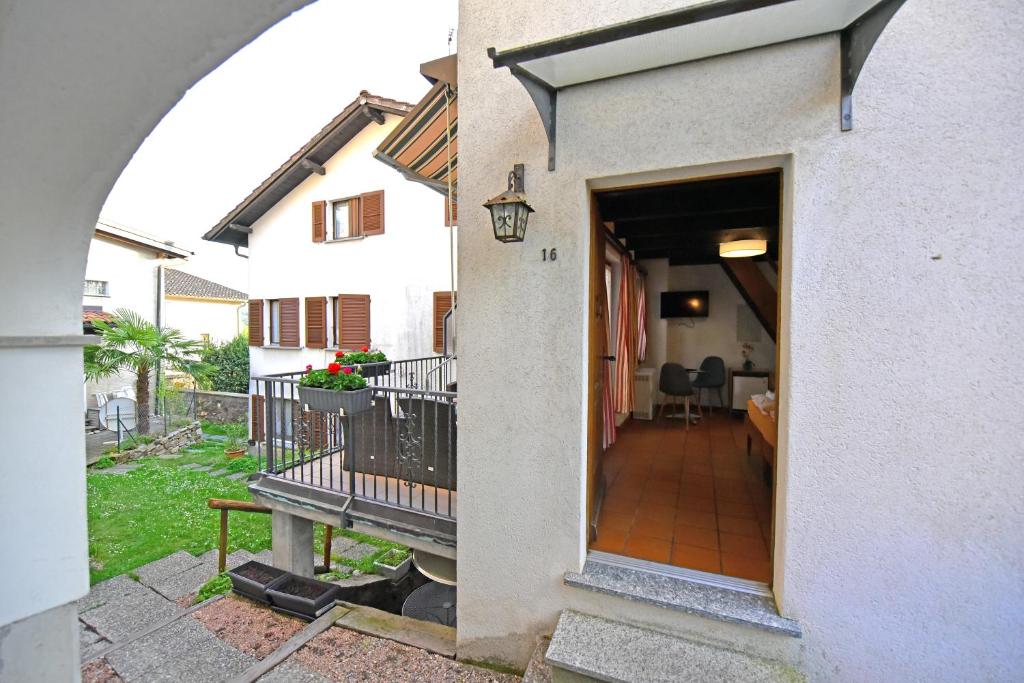 An entrance area is revealed under an arched overhang, leading to a stone pathway. A small balcony features potted flowers, while a grassy area is visible beyond. The building's exterior showcases a combination of windows and architectural details, contributing to the welcoming ambiance.
