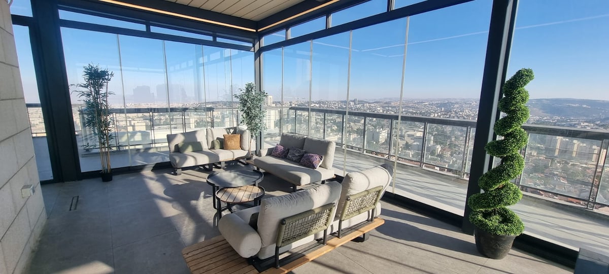 A glass-enclosed terrace features comfortable seating arrangements, providing stunning views of the cityscape. Soft cushions are arranged on the furniture, and greenery is introduced through potted plants, enhancing the space's tranquility. Large windows allow natural light to fill the area.