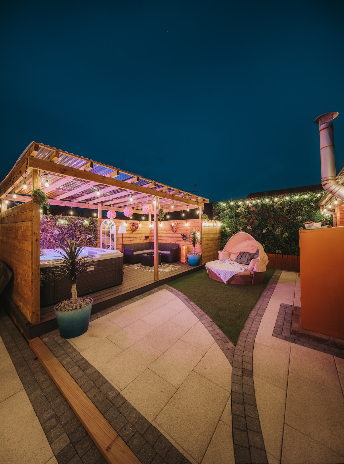 An outdoor seating area features a wooden gazebo with soft lighting, surrounded by lush greenery. Comfortable seating accommodates guests, while a cozy pink armchair enhances the tranquil atmosphere. A well-maintained path leads through the space, inviting relaxation under the evening sky.