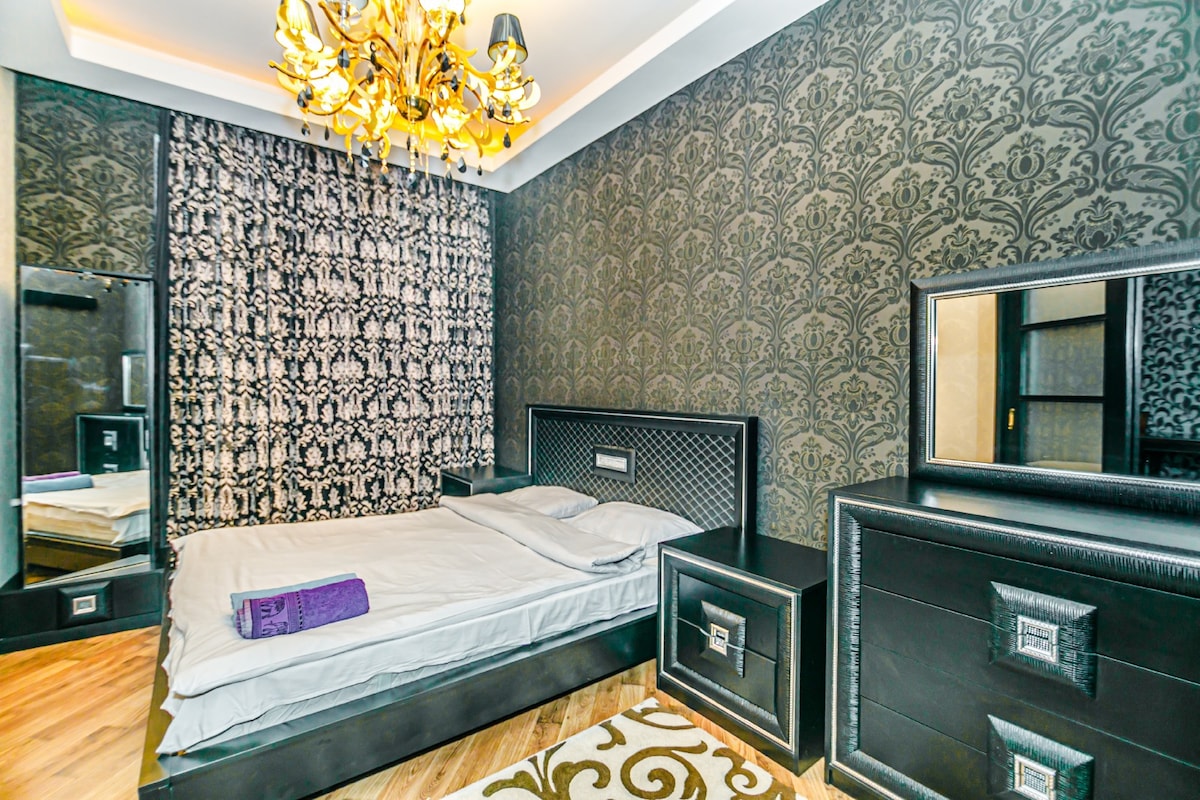 A bedroom is featured with dark, patterned wallpaper and a soft area rug. The spacious bed is positioned under a stylish chandelier, complemented by matching bedside tables and a large mirror that enhances the room's depth. A towel is neatly placed on the bed.