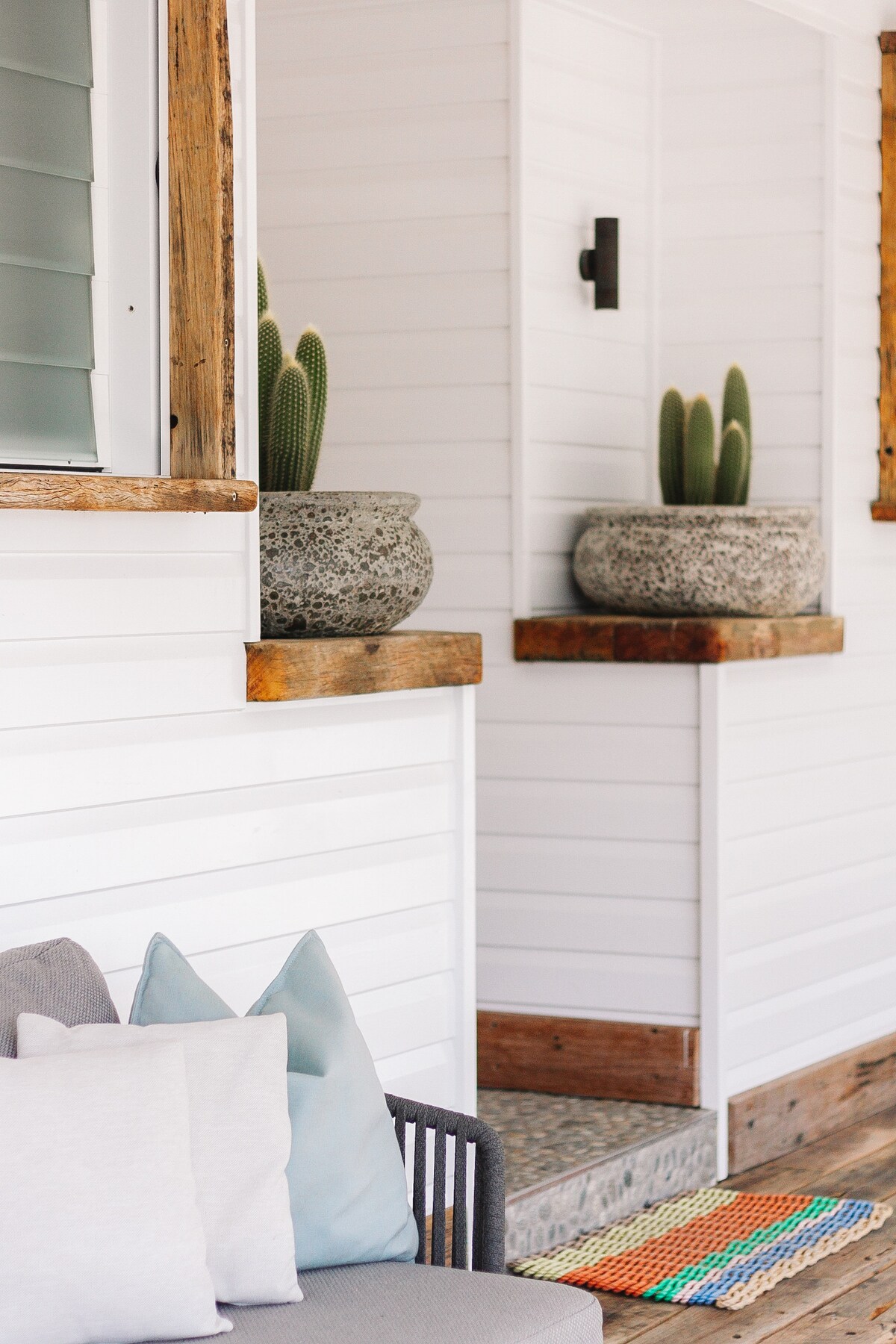 A welcoming entryway is presented with light-colored wooden accents and a spacious seating area featuring soft cushions. Decorative planters with cacti add a touch of greenery, while colorful woven mats provide a vibrant contrast to the white shiplap walls.