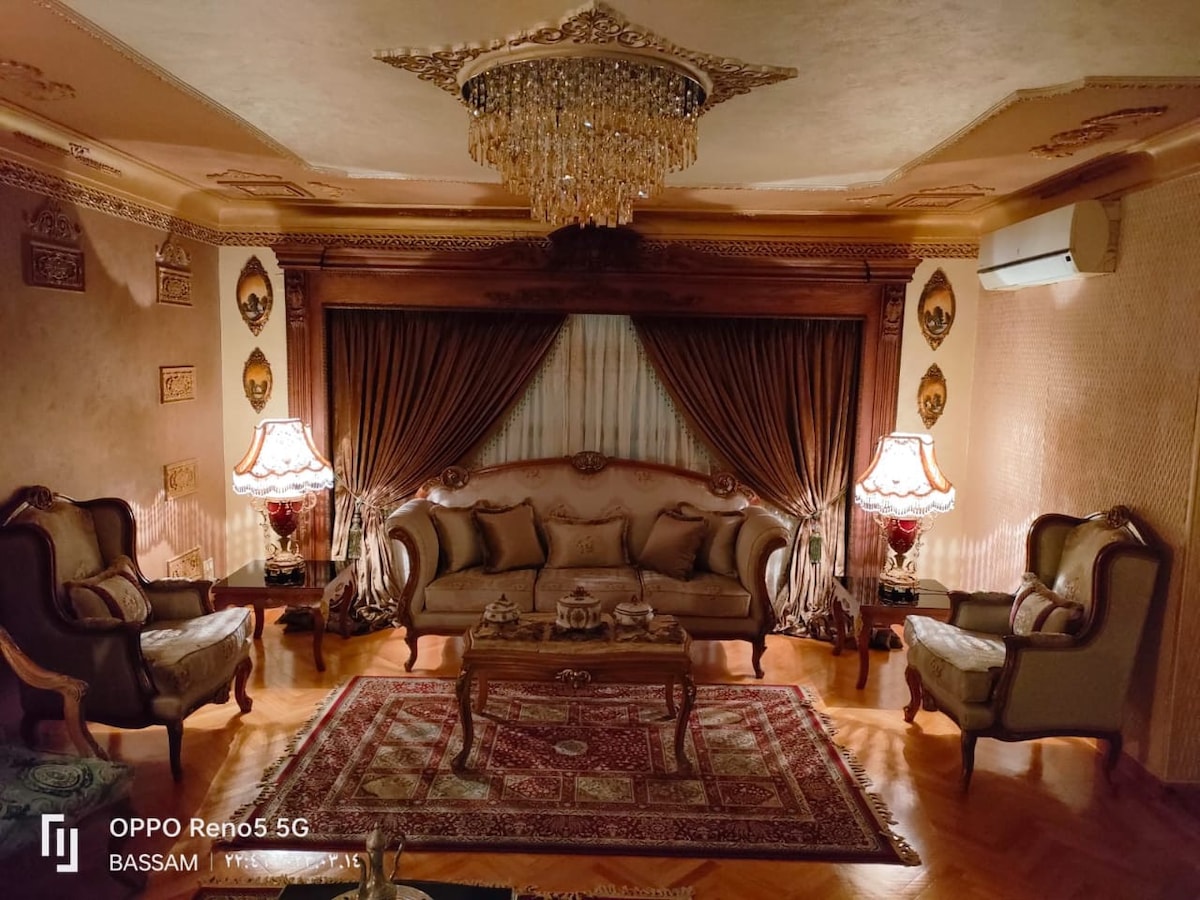 The elegantly furnished living room features a plush sofa set with ornate detailing, complemented by two matching armchairs. A decorative Persian rug covers the polished wooden floor beneath a crystal chandelier, which casts a warm glow over the richly textured walls and curtains.