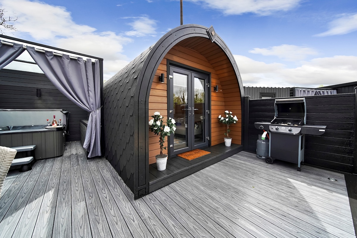 A modern glamping pod is showcased with a curved wooden exterior and glass doors. Two potted flowers flank the entrance, while a grill sits nearby. The spacious deck features comfortable seating and a hot tub, surrounded by tasteful outdoor decor.