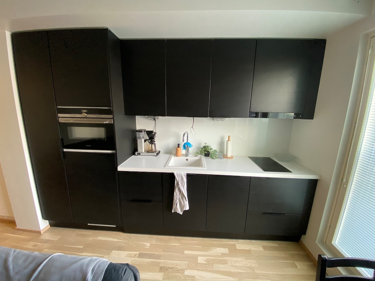 A modern kitchen features dark cabinetry and sleek countertops. A sink and a stove are positioned between the cabinetry. Appliances, including a coffee maker and oven, are integrated into the design. Natural light filters through windows, enhancing the functional layout.