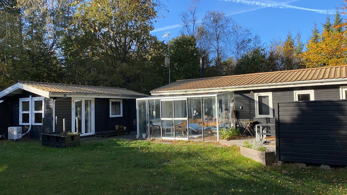 A charming summer house is shown, featuring a modern glass-enclosed dining area connected to a dark wood exterior. Lush green grass surrounds the structure, and clusters of trees are visible in the background, contributing to the peaceful setting.