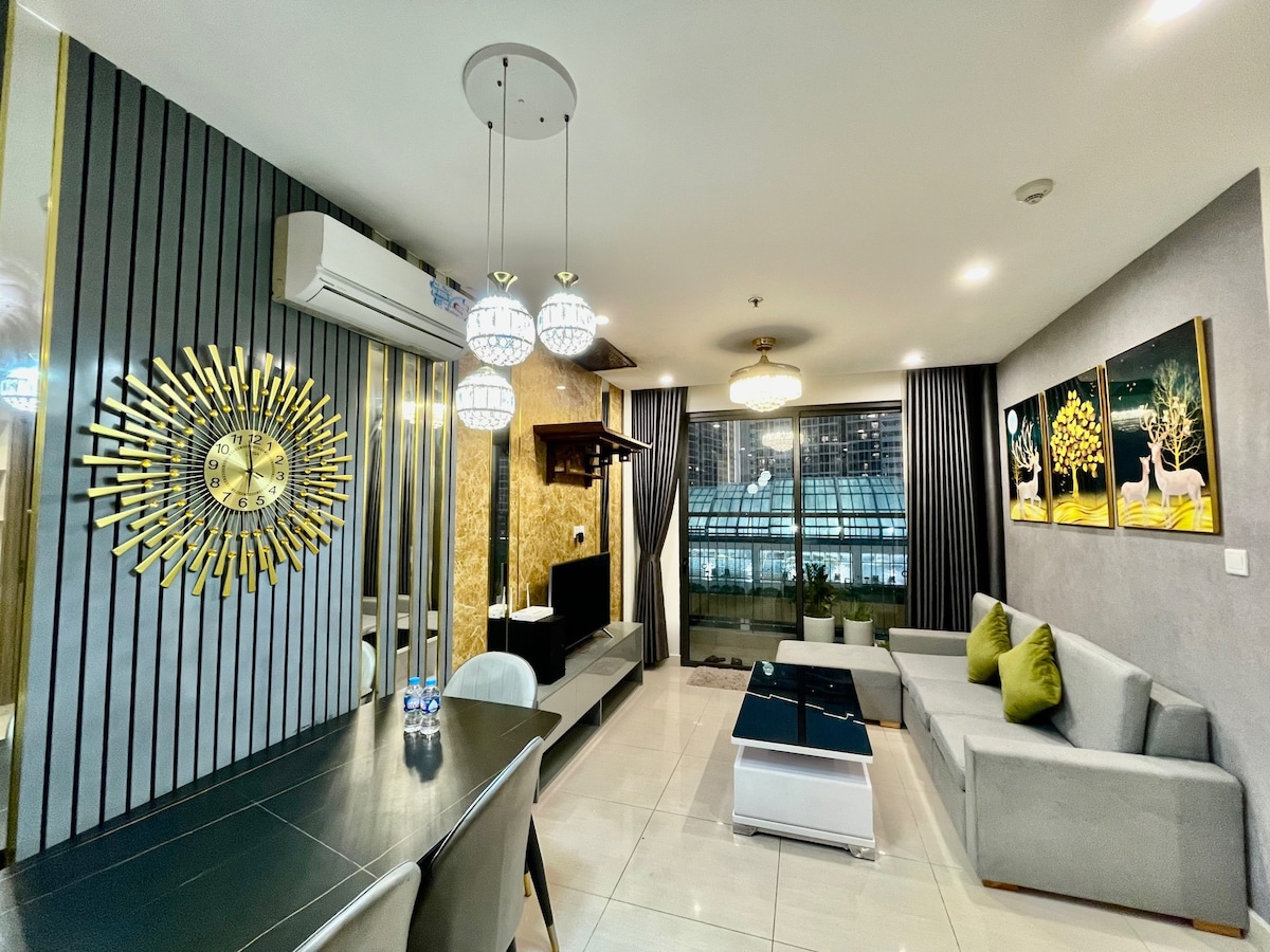 A modern living space features sleek furniture, including a gray sectional sofa and a coffee table, complemented by a dining area with a dark table. Decorative wall art and a gold-patterned accent wall add character, while large windows allow natural light to fill the room.