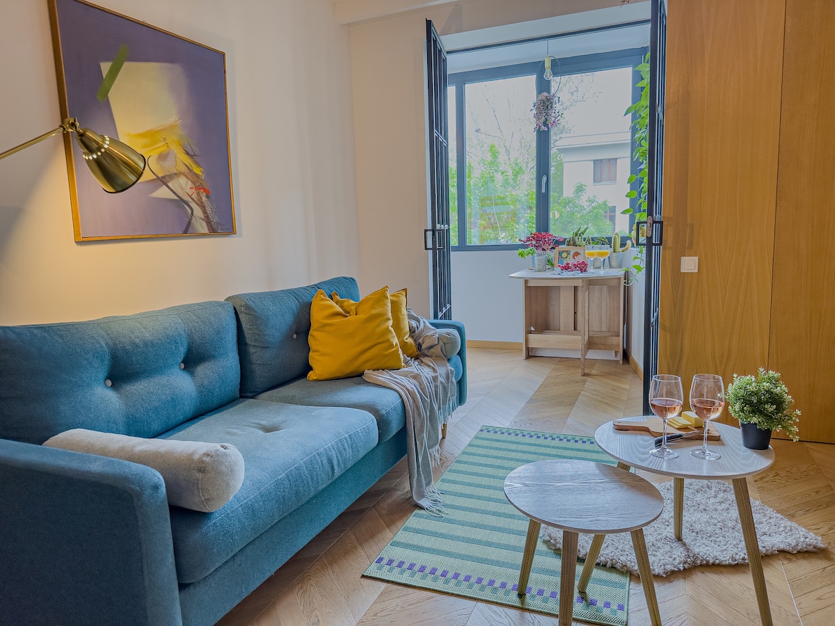 A cozy living area features a blue couch adorned with a yellow throw pillow. A small table holds two glasses. Bright natural light enters through open double doors, revealing a balcony with green foliage and a small dining setup.