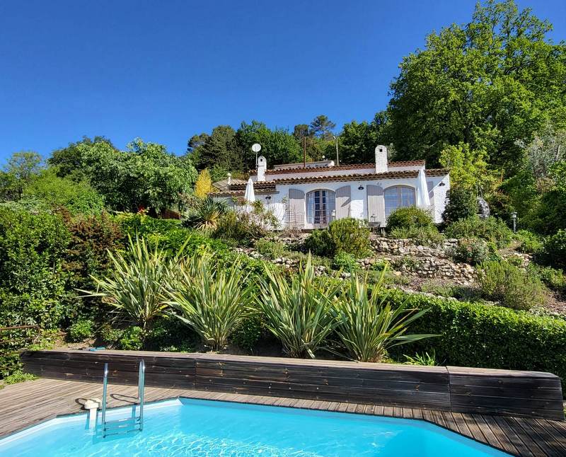 A charming white villa is set against a lush hillside, surrounded by well-maintained greenery and vibrant plants. A serene swimming pool is visible in the foreground, with wooden decking complementing the natural surroundings. Clear blue skies create a bright and inviting setting.