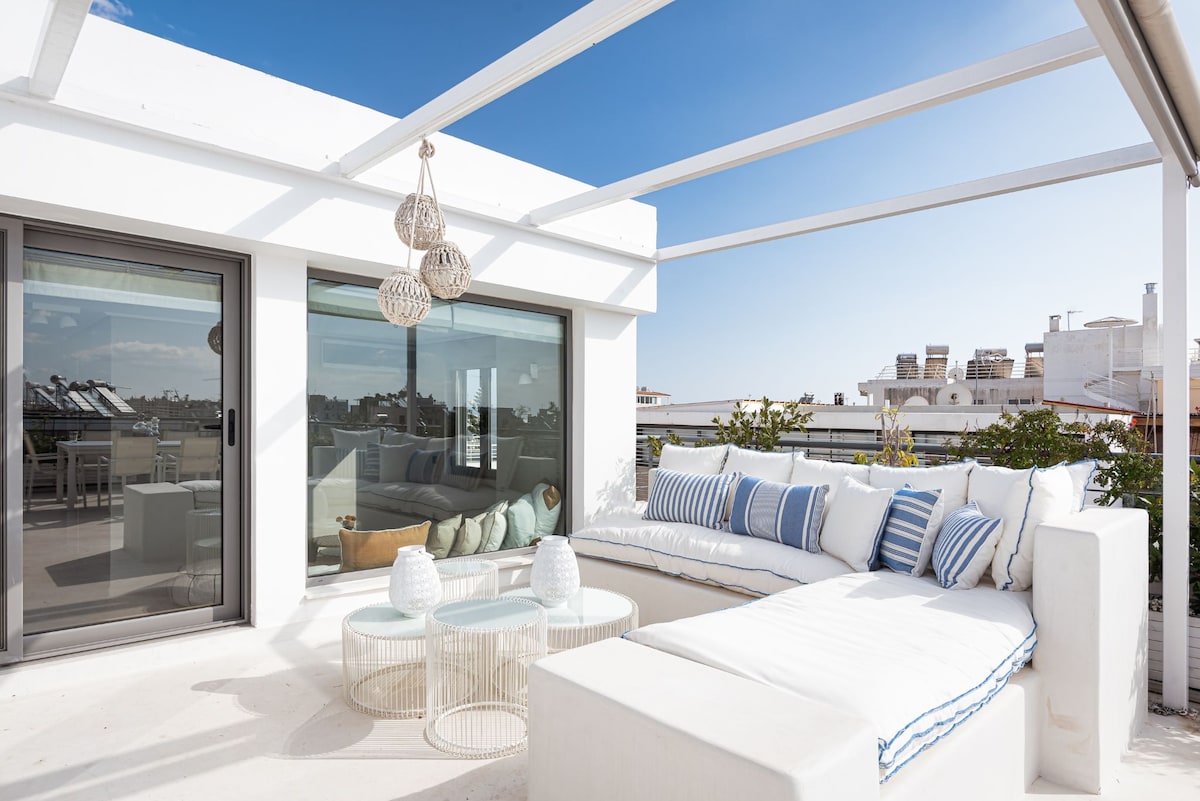A spacious terrace is showcased, featuring a comfortable seating area with plush cushions and decorative striped pillows. Light filters through the open space, enhancing the white decor. A glass table is positioned in the center, accompanied by woven pendant lights overhead.