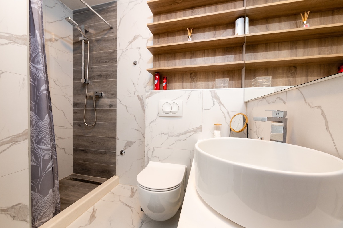 A modern bathroom features a sleek, curving sink atop a counter. A walk-in shower with geometric tile accents is visible, alongside a toilet and minimalist shelving displaying toiletries. Neutral tones and clean lines contribute to a contemporary aesthetic.