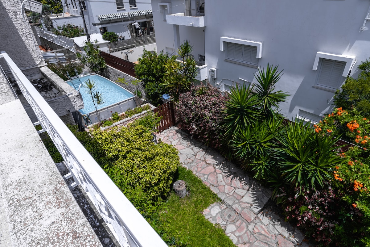 A serene outdoor area showcases a well-maintained garden featuring a variety of green plants and colorful flowers. A stone pathway winds through the garden, leading towards a swimming pool surrounded by shrubs and trees, providing a peaceful retreat.