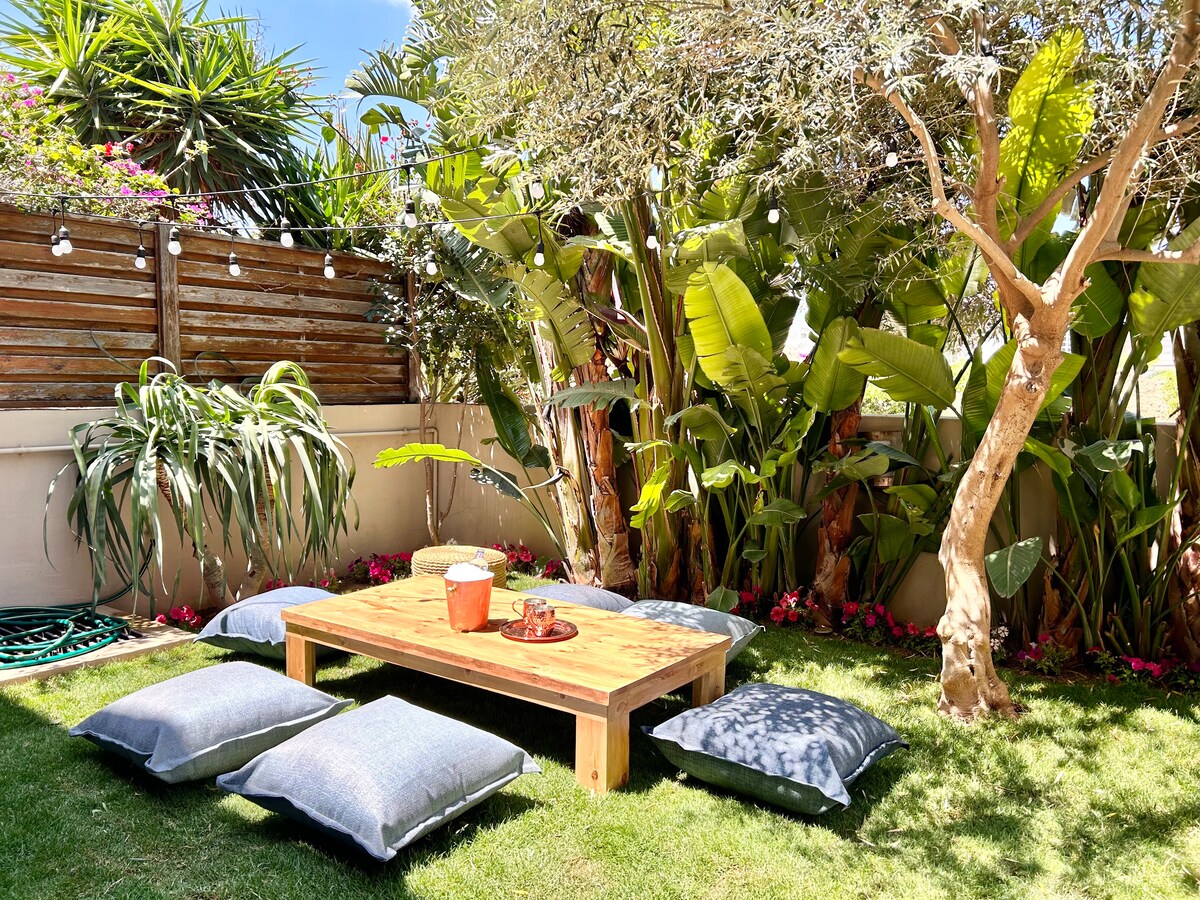 A serene outdoor space is surrounded by lush tropical plants, featuring a wooden table at the center. Cushions in various shades are arranged on the grass for seating, creating a comfortable area for relaxation or socializing. Soft lighting fixtures are visible above.