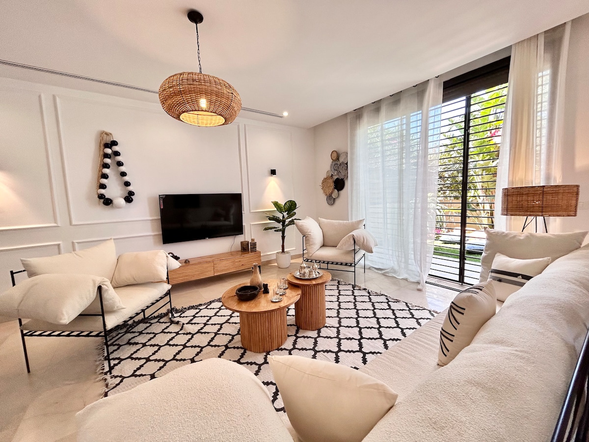 The living area showcases a light-filled space with a comfortable seating arrangement. Two round wooden tables are positioned between cozy armchairs. A large window features sheer curtains, allowing natural light to enter, while decorative elements add a modern touch to the atmosphere.