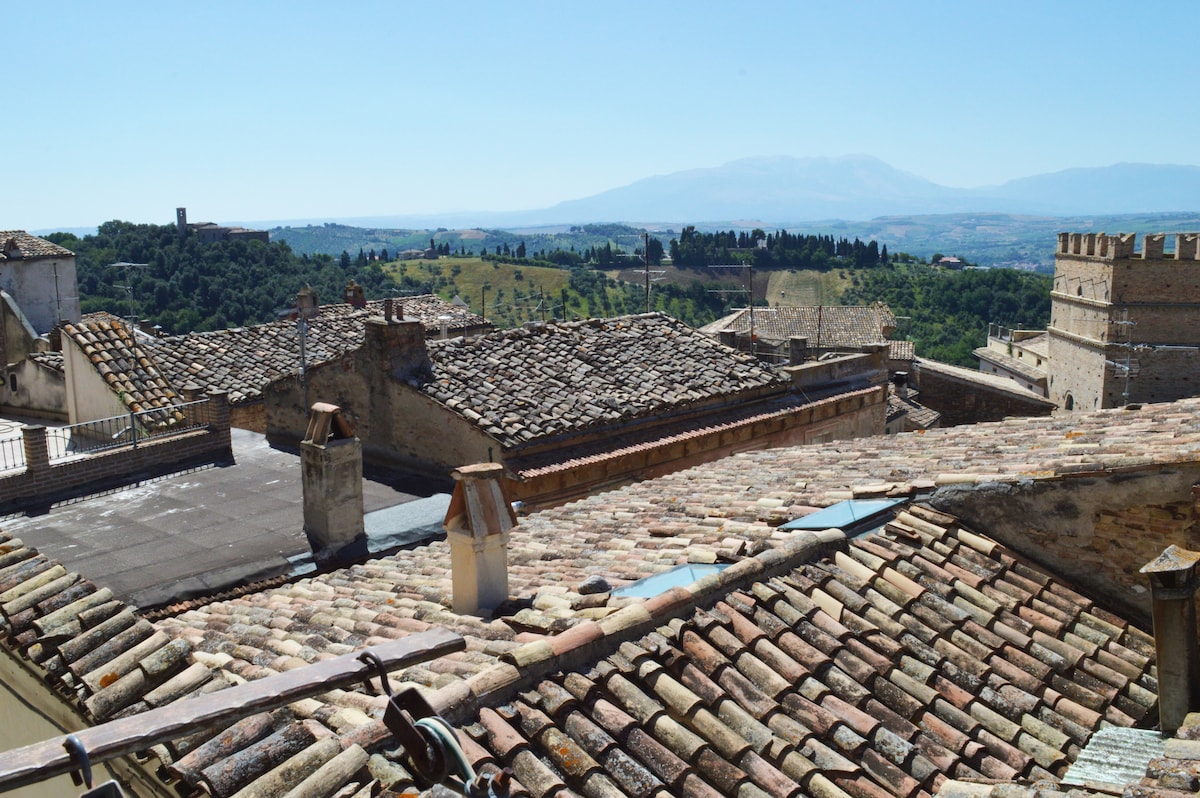 A scenic view of terracotta rooftops blending seamlessly into the rolling hills. The distant landscape reveals lush greenery, with hints of historical architecture visible amid the rooftops. A clear blue sky enhances the picturesque surroundings.