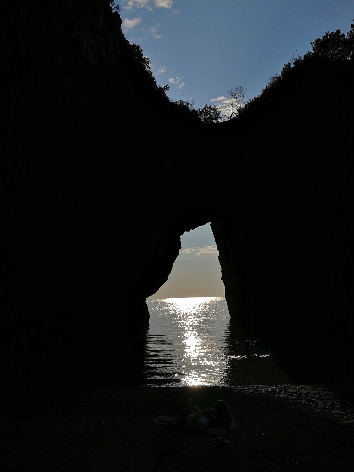 A silhouette of a natural arch stands against a tranquil sea, with sunlight reflecting off the water's surface. The surrounding cliffs rise steeply, framed by greenery. In the foreground, two figures are seated on the beach, enjoying the scenic view.