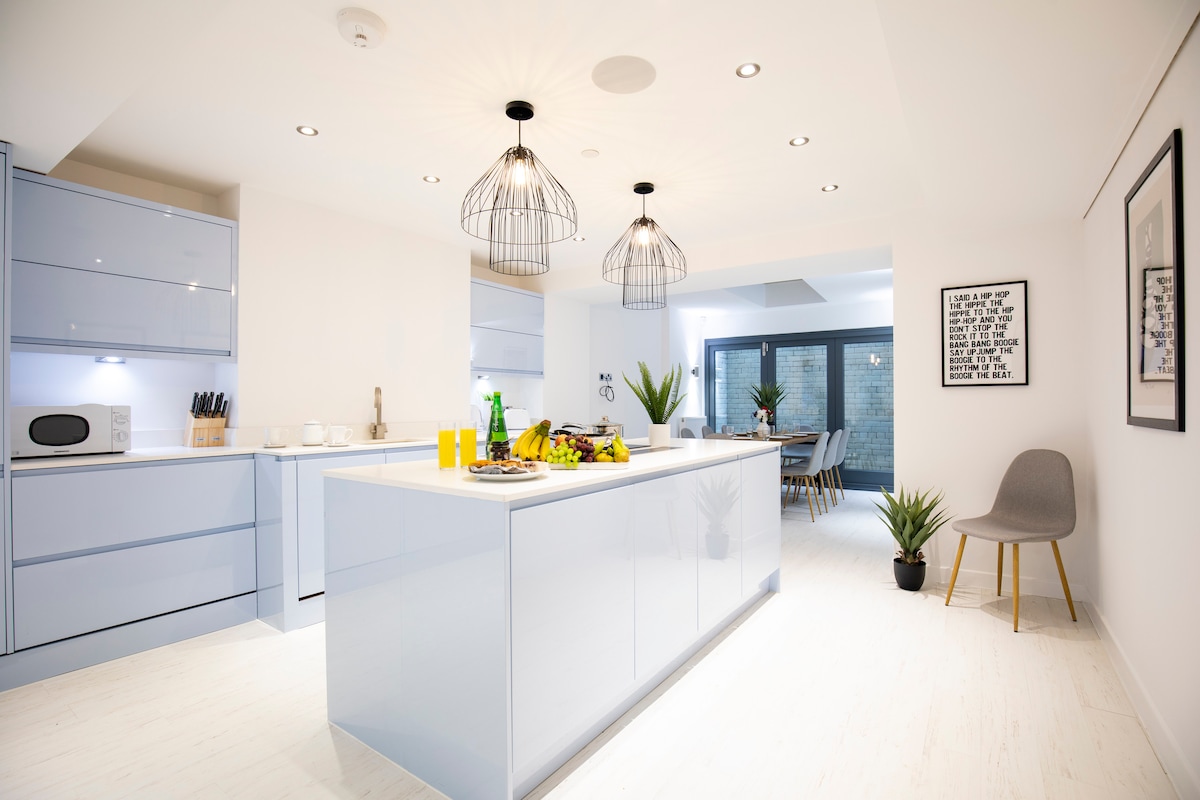 A modern kitchen layout features sleek blue cabinetry and a central island with built-in cooking area. Well-lit by decorative pendant lights, the space is accented by fresh greenery and a dining area visible through a glass door leading to an outdoor space.