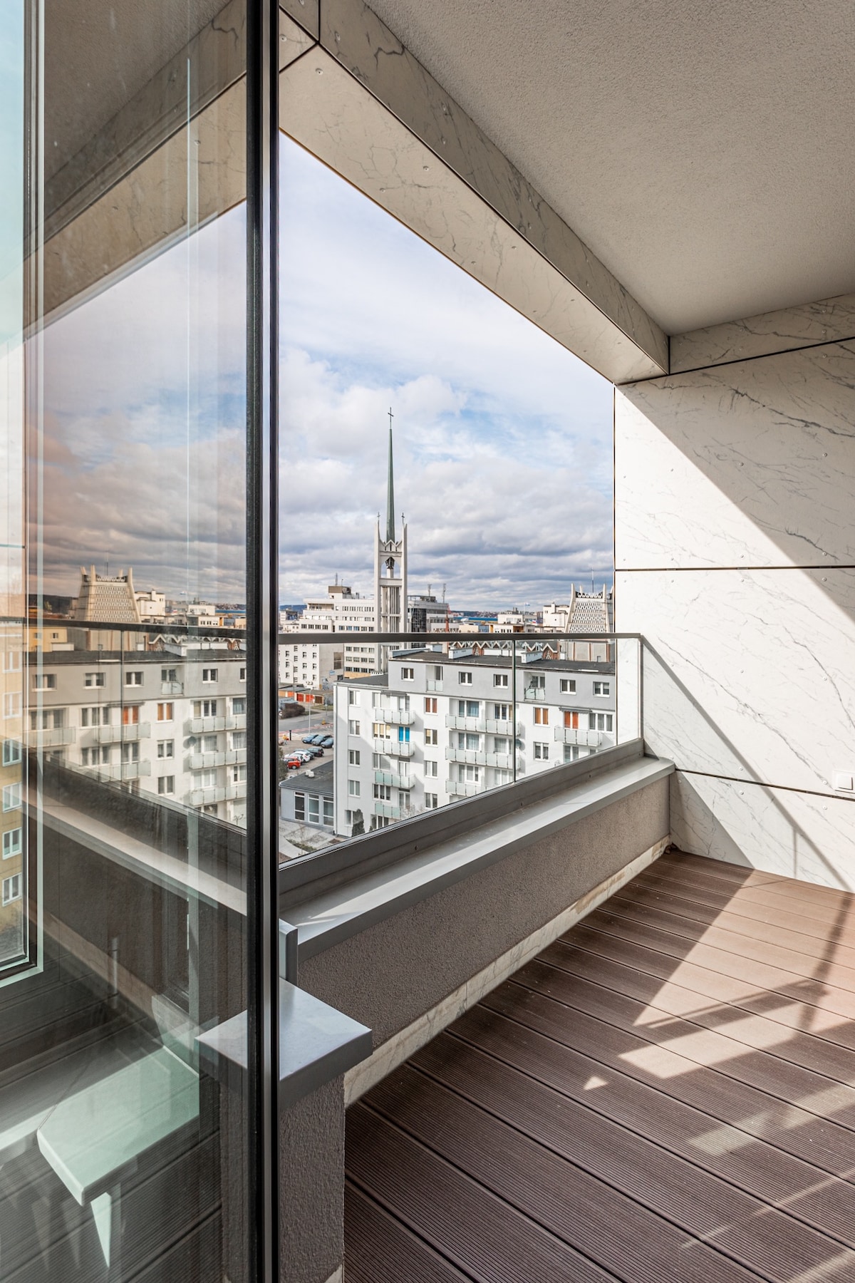 A modern balcony is depicted, showcasing a panoramic view of the city skyline, with a prominent tower spire in the distance. The design features sleek glass panels and clean lines, providing a sense of openness and connection to the outdoors.